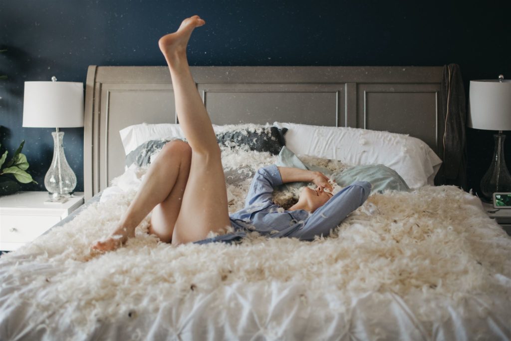 A woman in blue pajamas laying in a bed covered in feathers during a playful boudoir session in Chicago. 