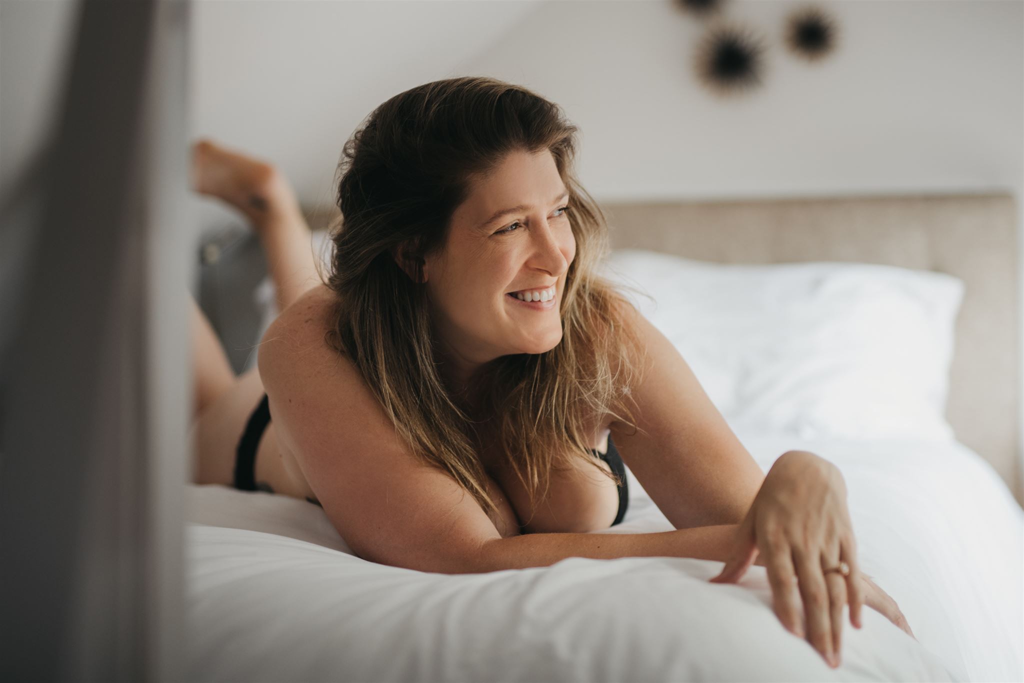 A woman in black lingerie lying on a bed with white sheets smiles during an inclusive boudoir photography session in Chicago with NSFW Boudoir.