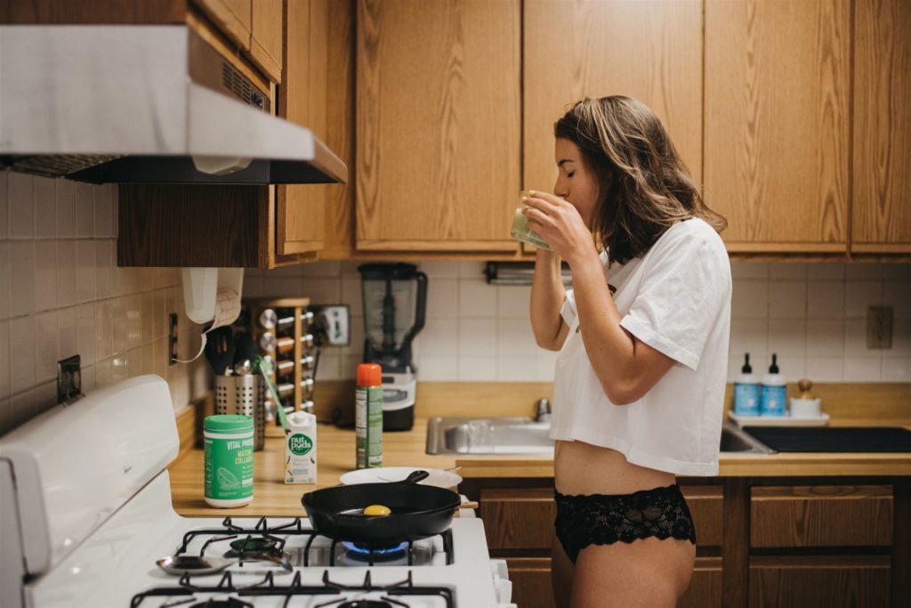 A woman in a t-shirt and lacy black underwear sipping coffee while cooking eggs in her kitchen during a lifestyle boudoir photo shoot in Chicago.