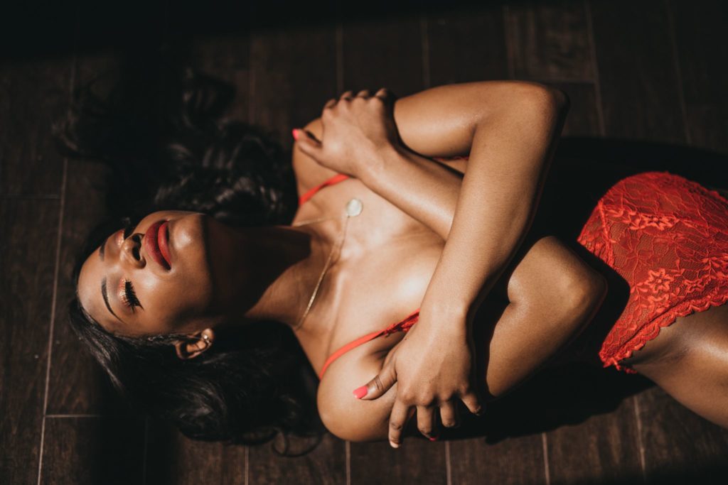 A woman wearing red lingerie lying on a wood floor with her arms wrapped around herself during a boudoir shoot in Chicago.
